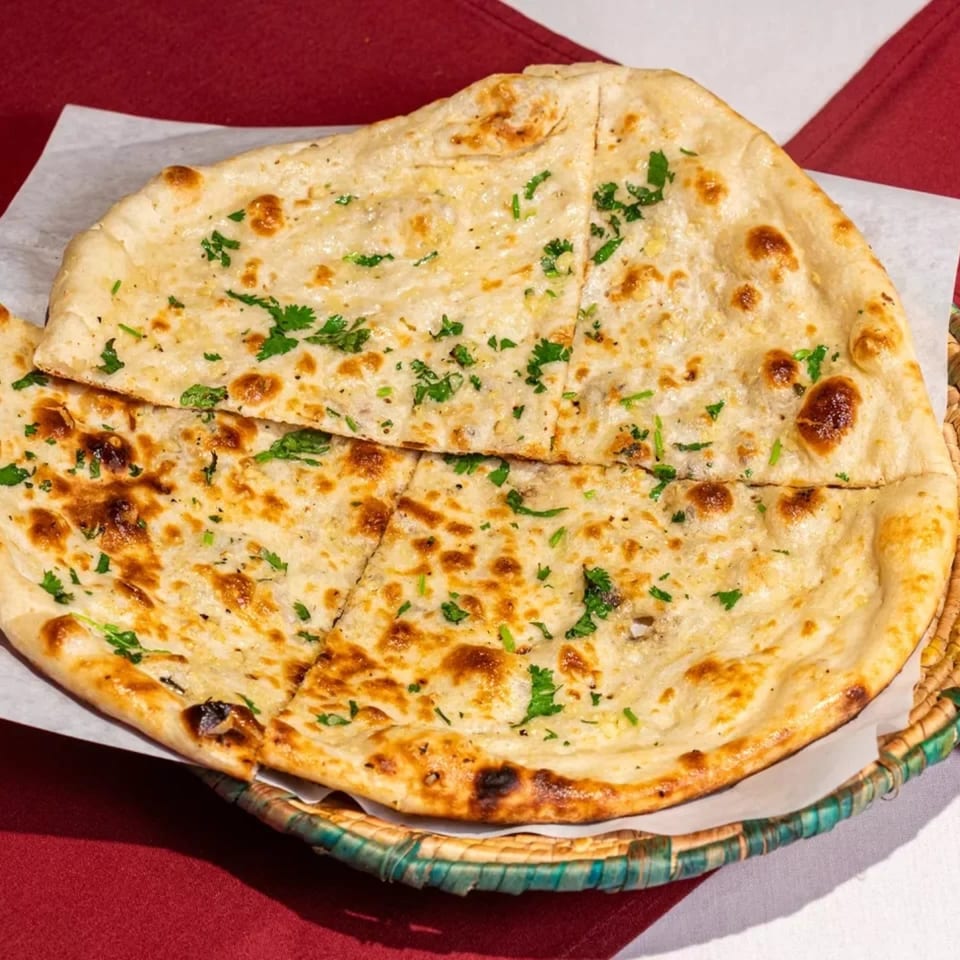 Golden-brown naan bread, garnished with fresh cilantro, served in a woven basket on a red and white tablecloth.