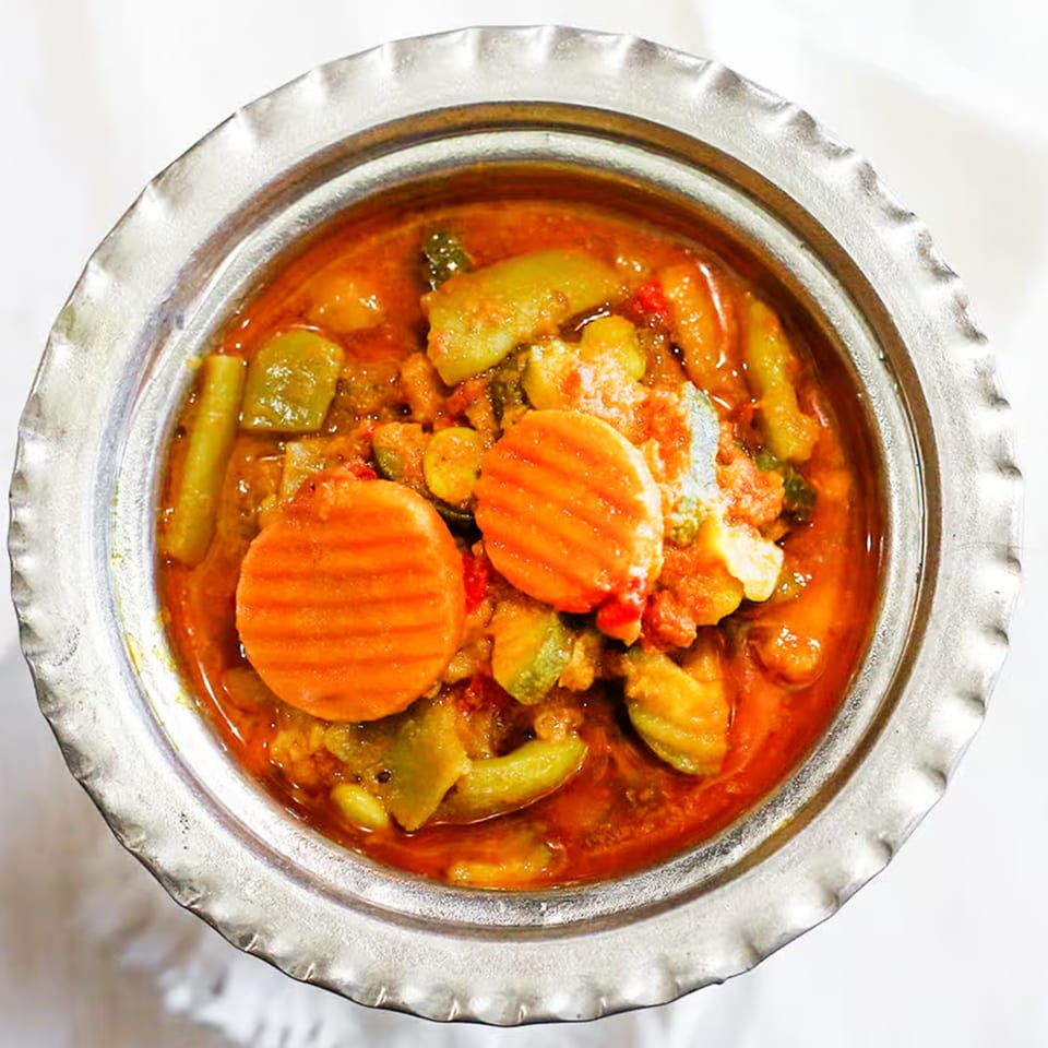 A close-up of a silver bowl filled with a vibrant Indian vegetable curry, featuring carrots and green beans.