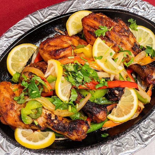 Sizzling Indian chicken tikka with bell peppers, onions, and lemon slices on a black platter.